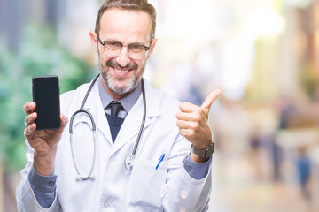 Middle age senior hoary doctor man showing smartphone screen over isolated background happy with big smile doing ok sign, thumb up with fingers, excellent sign