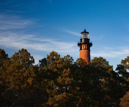 Lighthouse peeking above tall trees with a bright blue sky and light clouds in the background. Outer Banks, NC lighthouses attract tourists and history lovers. Room for text. Copy space top left.