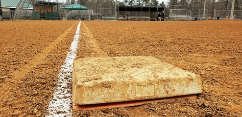 Dirty first base on a baseball field looking toward home and third base dugout.  Bright white foul line on clay diamond. Low angle. Little league baseball field in public park.