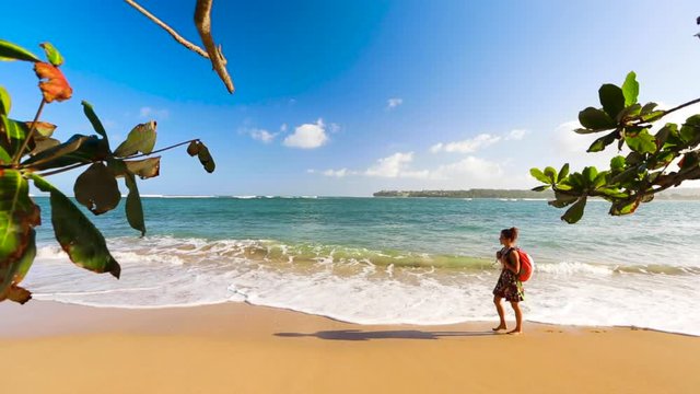 Tan woman wearing a backpack walks across Hawaii beach with ocean in the background with bright blue sky. Taking a walk on the shoreline with waves and beautiful sun.