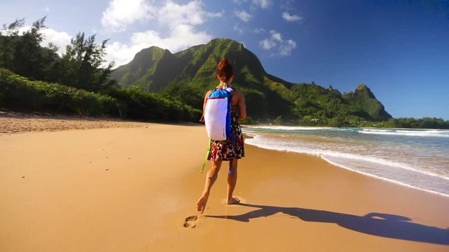 Tan woman wearing a backpack walks across Hawaii beach with ocean in the background with bright blue sky. Taking a walk on the shoreline with waves and beautiful sun.