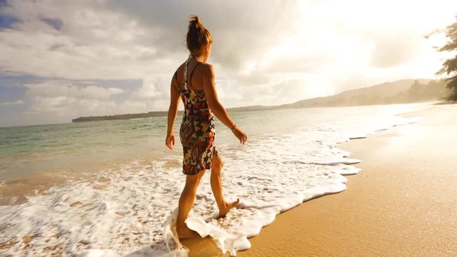 A woman walks across a beach in Hawaii with a sunset in the background and waves touching her feet. Tan girl on walking through the water leaving behind footprints.