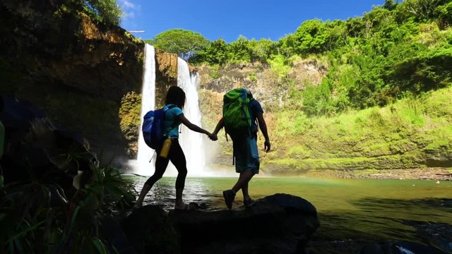 A couple silhouette in front of a waterfall in Hawaii. Romantic slow motion shot of couple holding hands by an ocean with backpacks on.