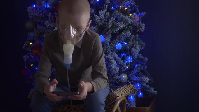Unhappy boy with shaved head inhales vapors containing medicines for inhaler to stop cough. Child is looking information about his illness in smartphone.  Respiratory medicine. Bronchitis problem
