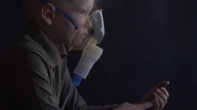 Unhappy boy with shaved head inhales vapors containing medicines for inhaler to stop cough. Child is looking information about his illness in smartphone.  Respiratory medicine. Bronchitis problem