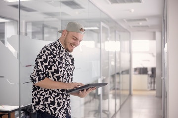 one young man, standing 20-29 years old, casual clothes, smiling laughing, using computer tablet. modern bright white office space.