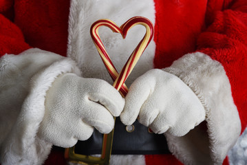 Closeup of Santa Claus hands holding two old fashioned candy canes forming a heart shape in front of his torso.