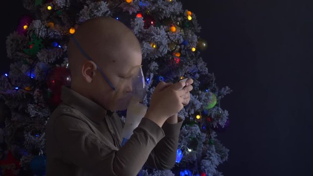 Unhappy boy with shaved head inhales vapors containing medicines for inhaler to stop cough. Child is looking information about his illness in smartphone.  Respiratory medicine. Bronchitis problem