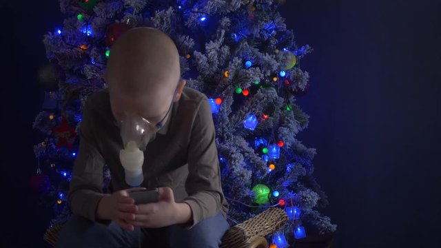 Unhappy boy with shaved head inhales vapors containing medicines for inhaler to stop cough. Child is looking information about his illness in smartphone.  Respiratory medicine. Bronchitis problem
