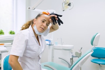 Tired woman dentist, background dental office chair. Female dentist taking off gloves wiping her...
