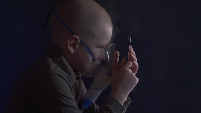 Unhappy boy with shaved head inhales vapors containing medicines for inhaler to stop cough. Child is looking information about his illness in smartphone.  Respiratory medicine. Bronchitis problem