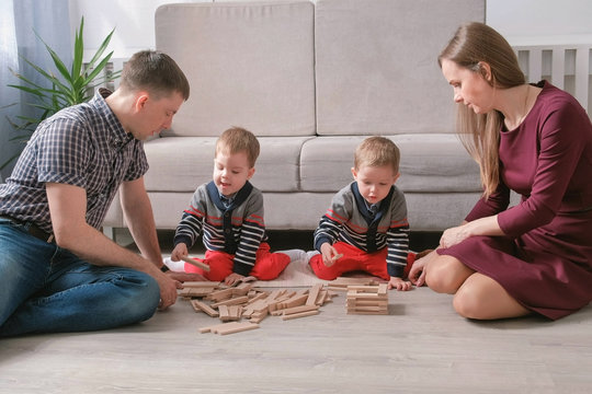 Family Mom, Dad And Two Twin Brothers Play Together Building Out Of Wooden Blocks On The Floor.