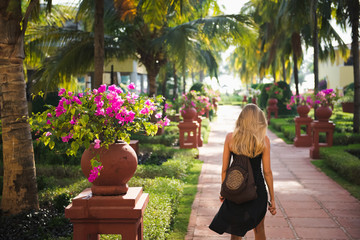 Blonde Woman in a Black Dress with a Backpack Walking Down a Hotel Path Surrounded by Lush...