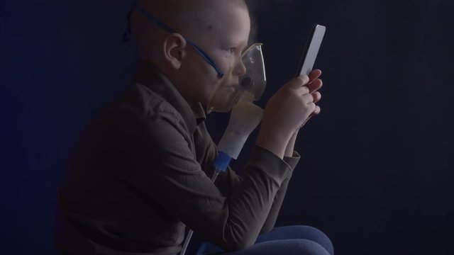 Unhappy boy with shaved head inhales vapors containing medicines for inhaler to stop cough. Child is looking information about his illness in smartphone.  Respiratory medicine. Bronchitis problem