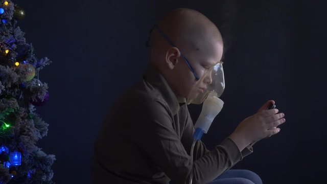 Unhappy boy with shaved head inhales vapors containing medicines for inhaler to stop cough. Child is looking information about his illness in smartphone.  Respiratory medicine. Bronchitis problem