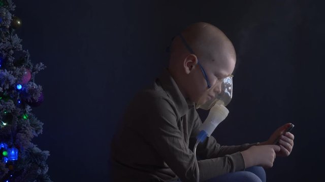 Unhappy boy with shaved head inhales vapors containing medicines for inhaler to stop cough. Child is looking information about his illness in smartphone.  Respiratory medicine. Bronchitis problem