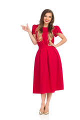 Elegant Woman In Red Dress And Gold High Heels Is Looking At Camera, Smiling And Pointing