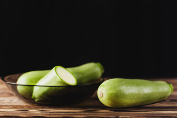 One whole zucchini near fresh zucchini in dark glass bowl on old brown wooden table on black background with copy space