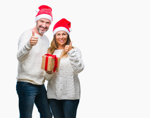 Middle age hispanic couple wearing christmas hat and holding gift over isolated background happy with big smile doing ok sign, thumb up with fingers, excellent sign