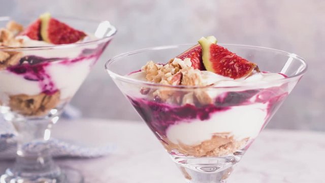 Figs pudding parfait with yogurt, blueberry jam, figs, hazelnut, and cookies in glass on white marble background. Flatlay top view.