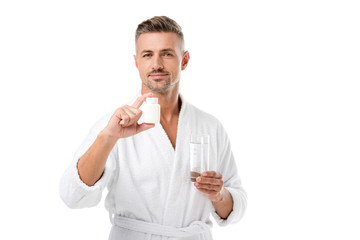 joyful adult man in bathrobe showing vitamins and holding glass of water isolated on white