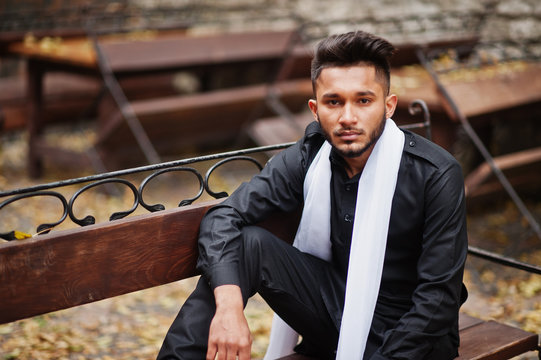 Indian stylish man in black traditional clothes with white scarf posed outdoor sitting on bench.