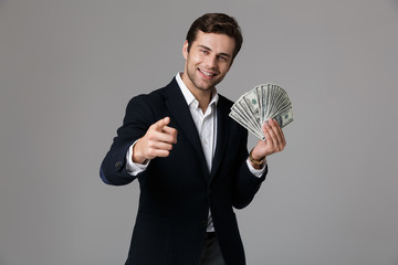 Image of successful businessman 30s in suit smiling and holding fan of money in dollar banknotes, isolated over gray background
