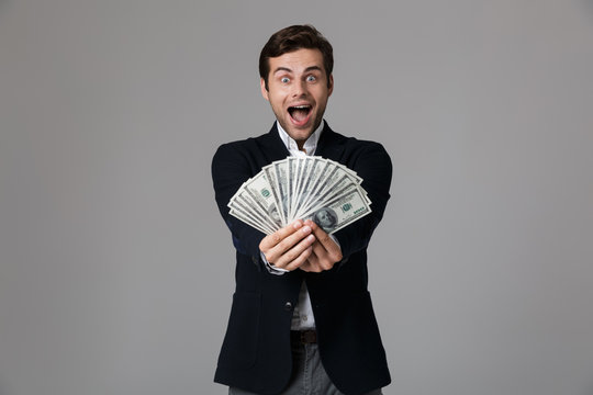 Image of joyful businessman 30s in suit smiling and holding fan of money in dollar banknotes, isolated over gray background