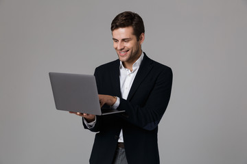 Image of pleased businessman 30s in suit rejoicing while using laptop, isolated over gray background