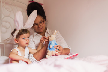 Obraz na płótnie Canvas Happy grandmother and her little grandson in the bunny ears sitting in a bed in the room at home and playing with a toy