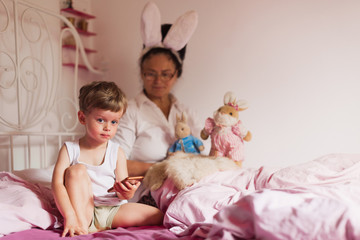 Obraz na płótnie Canvas Happy grandmother in the bunny ears sitting in a bed with the toys near her beloved little grandson who is holding a phone in hand in a bedroom
