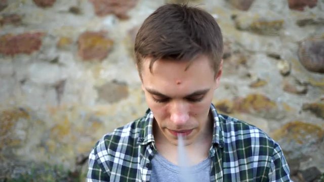 Beautiful model smokes electronic cigarette. Cute guy smiles and blows smoke from vaping. A man in a plaid shirt near the wall smokes an electronic cigarette. Handsome guy blowing smoke in the cell.