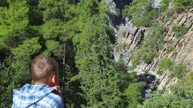 Back view of young white boy sitting on top of cliff of canyon resting and looking at beautiful mountains with green trees growing on rocks. Real time full hd video footage.