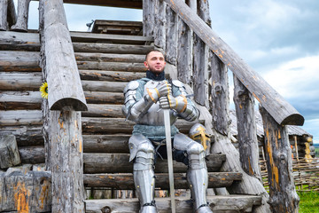 Knight in the armor on the wooden steps. Knightly armor and weapon. Semi - antique photo.