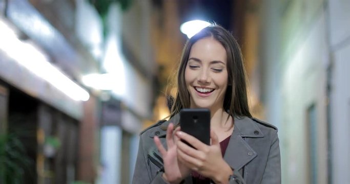 Front view of a happy woman walking towards camera using a smart phone in the night in the street