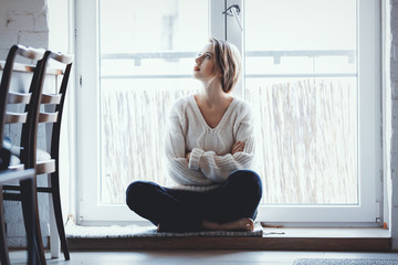 young girl dressed in a sweater and jeans is sitting at the threshold of the window at home