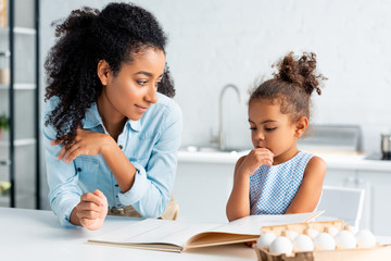 pensive african american mother and daughter choosing recipe from cookbook in kitchen