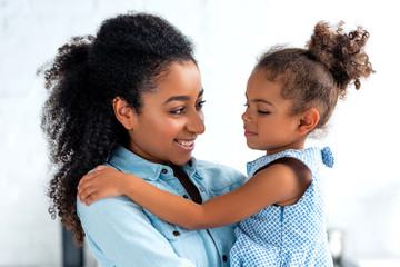 smiling african american mother and daughter hugging in kitchen and looking at each other