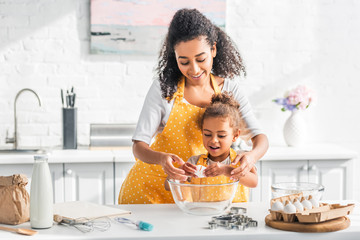 smiling african american mother helping daughter breaking egg for preparing dough in kitchen - Powered by Adobe