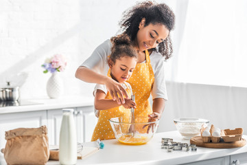 african american mother helping daughter whisking eggs for dough in kitchen