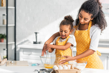 african american mother and daughter in yellow aprons whisking eggs for dough in kitchen