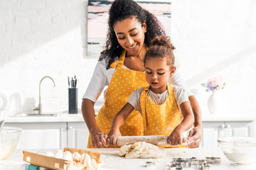 african american mother helping daughter rolling dough with rolling pin in kitchen