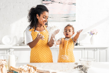 african american mother and daughter in aprons preparing dough and having fun with flour in kitchen