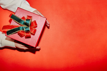 Fototapeta premium female hand in sweater holding a red present box for Christmas, New Year, Valentine'a day on red background, top view with copy space