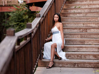 Beautiful brunette girl in a white dress sits on wooden stairs at the park