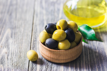 Extra vergin olive oil in glass jar, marinated olives in wooden bowl on rustic background. Selective focus, space for text.