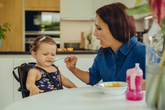 Mother helping disgusted baby daughter eating fruit pulp in kitchen