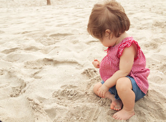 Nine month old baby girl sitting on the beach in beautiful summer day. Little pretty cute european infant child with dark hair in red plaid shirt and jeans shorts sitting on the sand of the beach.