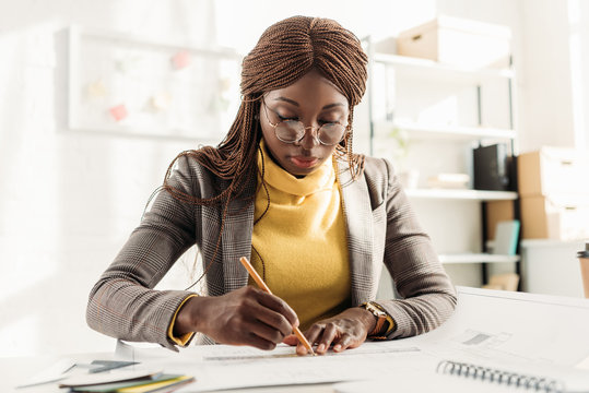 concentrated african american female architect in glasses holding pencil and working on project at desk with blueprints in office - Powered by Adobe