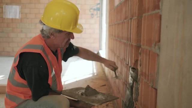 People working in construction site. Busy caucasian man at work in new house inside apartment building. White manual worker using equipment and tools to work on brick wall with cement